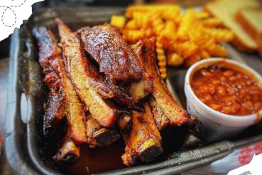 Succulent grilled pork ribs with crispy seasoned fries and baked beans in a small cup, served on a metal tray for a hearty American barbecue meal, close-up shot of delicious comfort food.