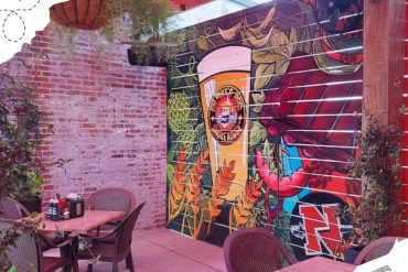 Vibrant outdoor restaurant patio with colorful mural featuring a beer glass, creative beer-themed artwork, and brick wall, perfect for casual dining and socializing.