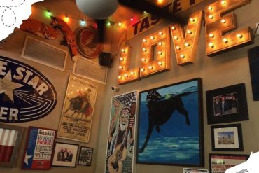 Bright indoor restaurant decor with large illuminated "LOVE" sign, colorful string lights, artistic wall art including cowboy and musical poster themes, creating a lively and inviting atmosphere.