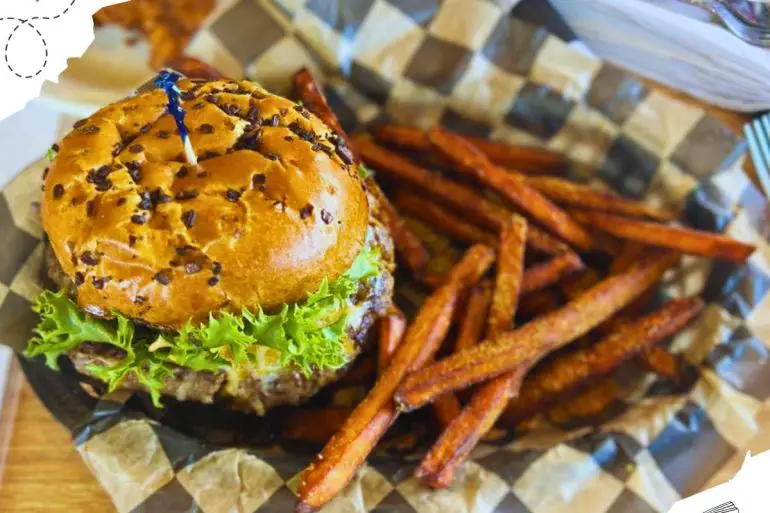 Burger with toothpick on top on blue and white checkered paper and sweet potato fries.