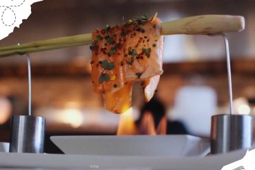 Seared salmon fillet with herbs and spices held over a flame on a skewer, ready for serving at a restaurant or culinary event.