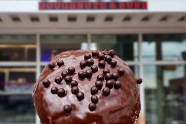 Chocolate-covered donut with chocolate sprinkles held in front of HERO Doughnut & Deli shop.
