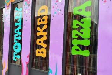 Colorful storefront window with vibrant signs reading "TOURIST," "BACKED," and "DAY 2," decorated with stars and dreamy pastel designs, promoting travel experiences and destinations.