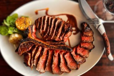 Juicy grilled steak served with fresh herbs, citrus, and drizzled with sauce, on a white plate perfect for savoring delicious American BBQ cuisine.