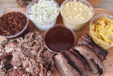 Juicy barbecue ribs with shredded pork, baked beans, potato salad, coleslaw, mac and cheese, and barbecue sauce on a wooden platter. Perfect for a Southern barbecue feast.