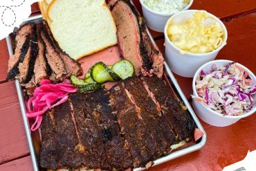 Smoked barbecue platter with ribs, brisket, sliced bread, pickles, and colorful sides including coleslaw and macaroni salad, served on a metal tray for a classic Southern dining experience.