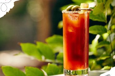 Vibrant Bloody Mary cocktail with green olives and celery on a sunny outdoor table, perfect for travel and leisure moments. Ideal for travelers seeking relaxing destinations with delicious drinks.