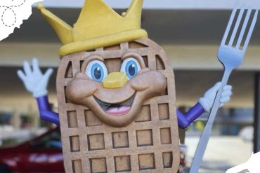 Waffle mascot character wearing a crown, holding a fork, cheerful expression, outdoor setting, playful and fun travel-themed image for destination exploration.