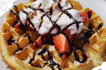 Buttermilk waffle topped with fresh strawberries, whipped cream, and drizzled with chocolate syrup, served on a white plate. Ideal for breakfast or brunch, highlighting delicious dessert options.
