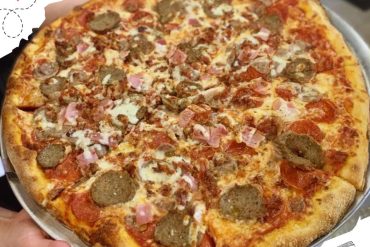 Savory pizza topped with sausage, ham, and mushrooms on a golden crust, served on a metal tray, perfect for travel or food lovers exploring diverse destinations.