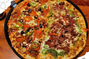 Savory pizza with mixed toppings including olives, peppers, and meat, served on a black pizza pan in a cozy restaurant setting.