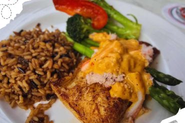 Grilled fish fillet with melted cheese, served with seasoned rice, fresh broccoli, asparagus, and roasted red pepper on a white plate for a delicious seafood meal.