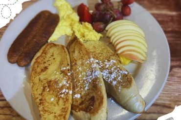 Golden French toast with powdered sugar, scrambled eggs, sausage links, fresh apple slices, and grapes on a white plate. A hearty breakfast dish popular for travel mornings and food tourism.