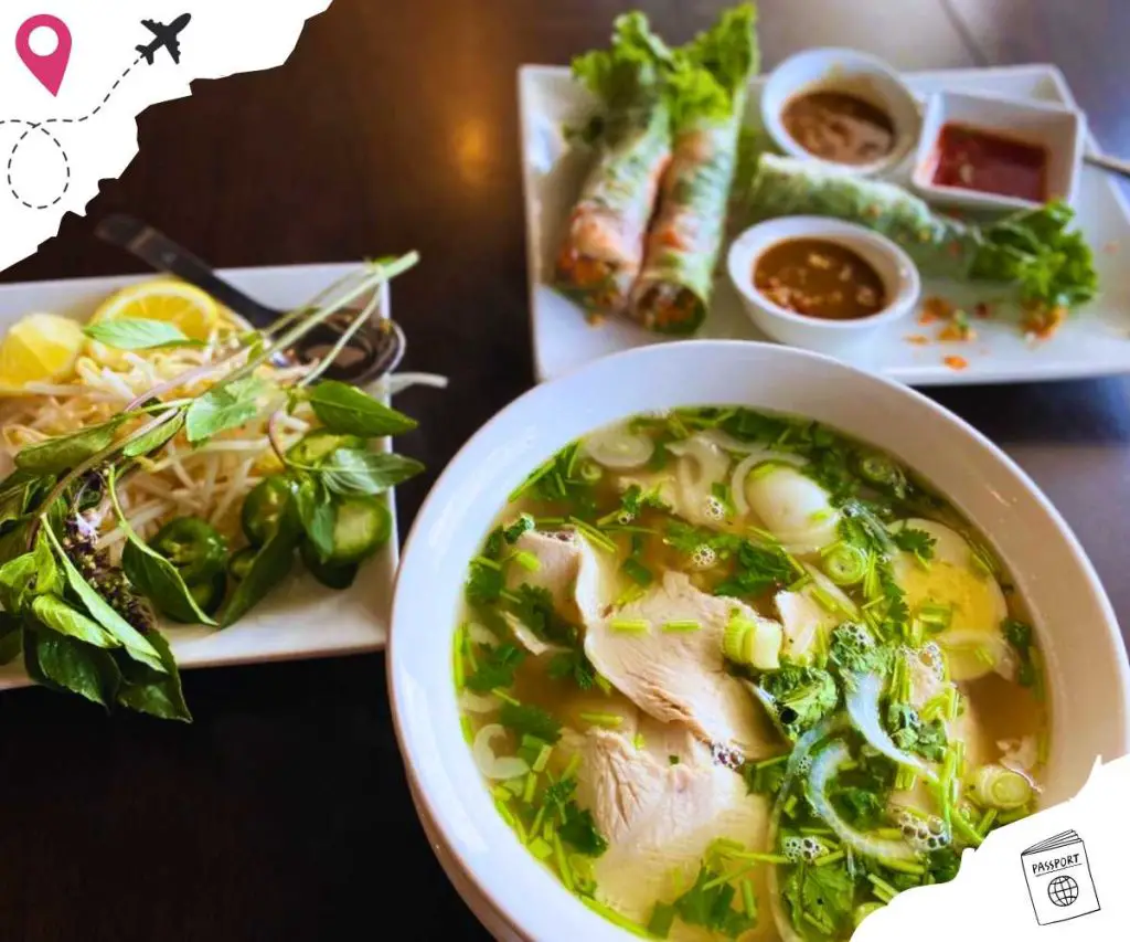 A bowl of pho, a plate of fresh vegetables, and a plate of spring rolls with a bowl of sauce on it.