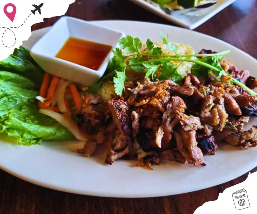 A plate of grilled pork garnished with cilantro and with vegetables and a small bowl of sauce on the side.