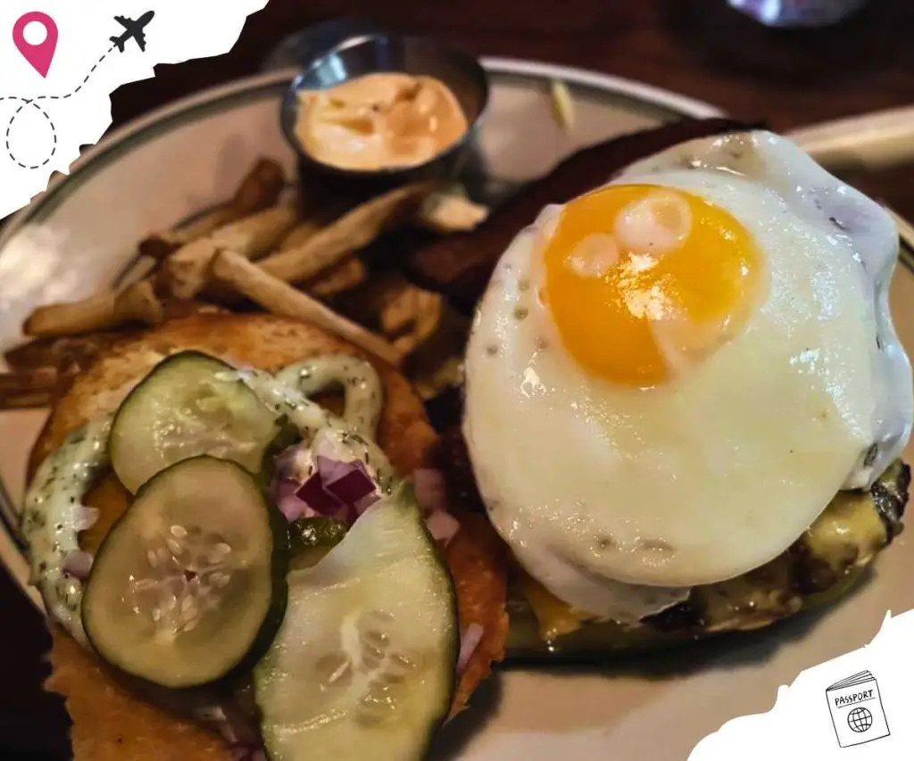 An opened burger with a sunny side up egg on the bottom piece, as well as French fries and a cup of dipping sauce on the side.