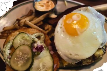 An opened burger with a sunny side up egg on the bottom piece, as well as French fries and a cup of dipping sauce on the side.