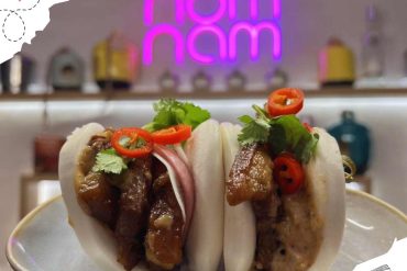 Beef bao bun dishes with fresh herbs and chili toppings at a restaurant in front of decorative neon sign, Asian cuisine, street food, gourmet food, trendy dining atmosphere.