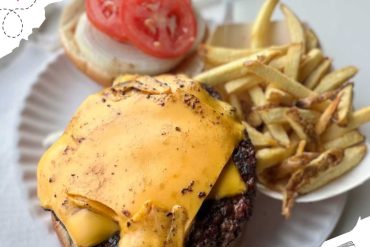 Juicy cheeseburger with melted cheddar cheese, fresh tomato slices, and a side of crispy French fries on white paper plate. Perfect for a delicious travel-inspired meal.