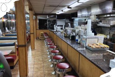 Bright diner interior with a long counter and red cushioned stools, featuring an open kitchen with plates and baking trays, perfect for a classic American lunch or casual meal.