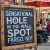 Chalkboard sign advertising a "Sensational Hole in the Wall Spot Fargo, ND" restaurant, with outdoor seating and colorful umbrellas, capturing a lively dining scene perfect for travel and food destination SEO.