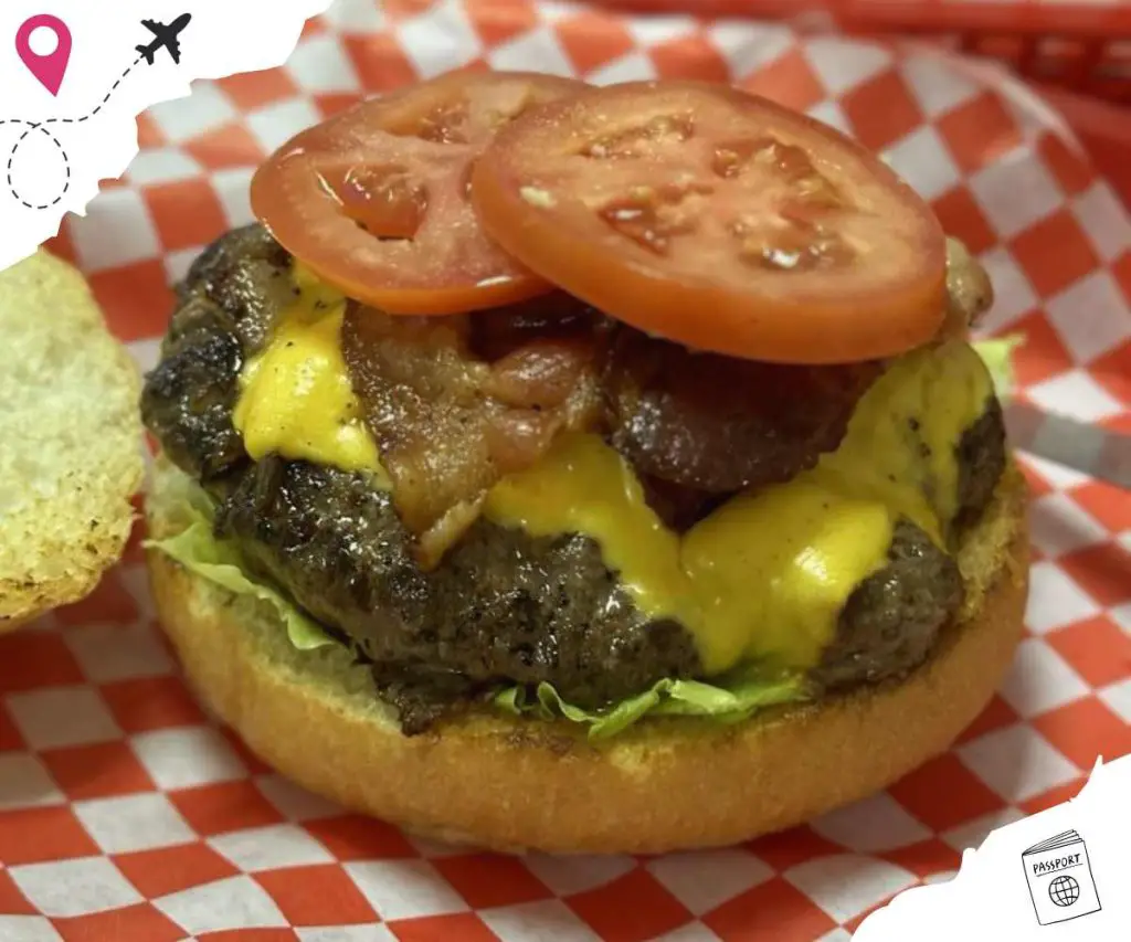 Juicy cheeseburger with melted cheese, beef patty, fresh lettuce, tomato slices, and sesame seed bun on checkered red and white paper background.