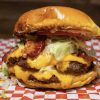 Juicy cheeseburger with bacon, lettuce, tomato, and melted cheddar cheese on a toasted bun, served on red checkered paper for a classic fast-food dining experience.