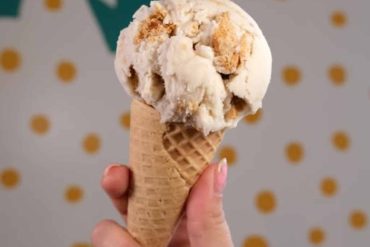 Creamy vanilla and caramel ice cream scoop in a waffle cone, held in a person's hand against a colorful polka dot background. Perfect for ice cream lovers and dessert enthusiasts.