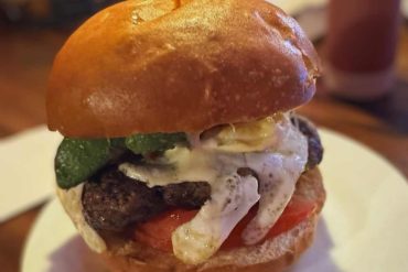 Succulent cheeseburger with melted cheese, fresh tomato, pickles, and a toasted bun, served on a white plate at a restaurant, showcasing American-style comfort food and gourmet burger dining experiences.