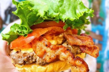 Freshly prepared gourmet burger with lettuce, tomato, crispy bacon, and melted cheese, served in a soft bun. Perfect for a tasty outdoor meal and travel food adventures.