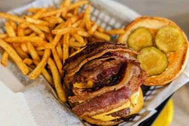 Crispy bacon cheeseburger with pickles and fries, fast food meal, American comfort food, savory burger, sandwich meal, casual dining, tasty burger, fries on paper basket, classic American cuisine, delicious fast food.