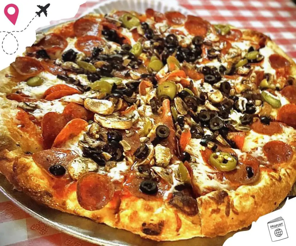 Slice of loaded pizza with pepperoni, black olives, green olives, mushrooms, and peppers on a metal tray. Perfect for travel food lovers, representing classic comfort cuisine.