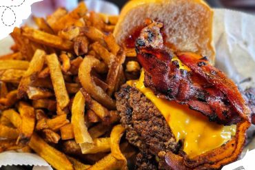 Crispy bacon cheeseburger with melted cheddar cheese, seasoned beef patty, and fresh bun served with crispy French fries on side at a restaurant.
