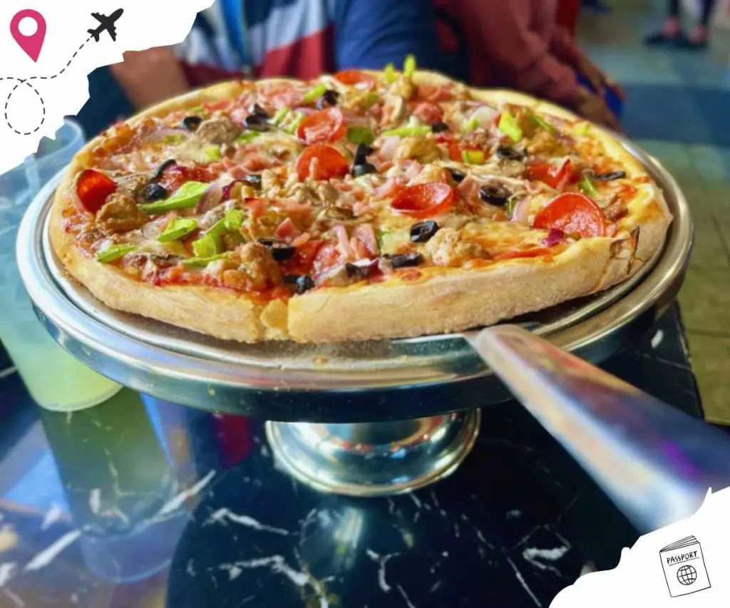 Freshly baked pizza with assorted toppings including pepperoni, olives, tomatoes, and green peppers on a stainless steel serving tray. Perfect for travel food adventures and international cuisine experiences.