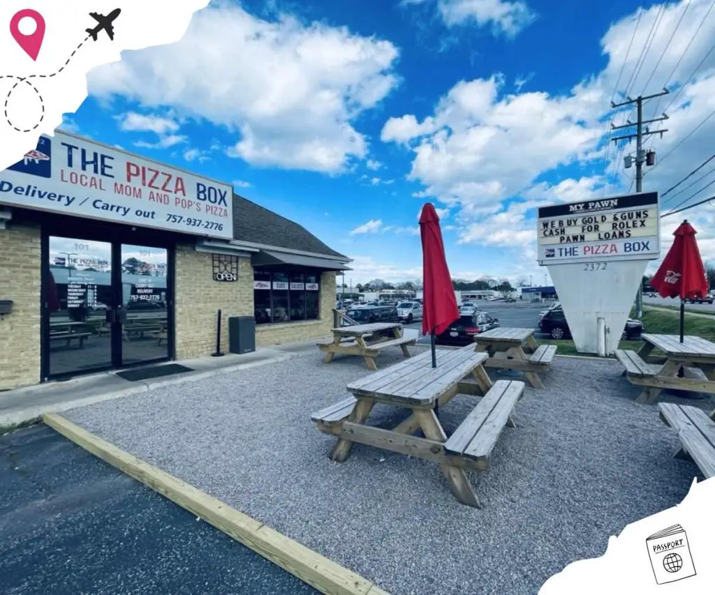Pizza restaurant storefront with outdoor seating, wooden picnic tables, red umbrellas, bright blue sky with clouds, and signage for local pizza, pawn shop, and cash loan services.