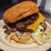 Juicy cheeseburger with cheddar cheese, crispy bacon, seasoned beef patty, fresh lettuce, and toasted bun on a disposable plate. Perfect comfort food from Go To Destinations.