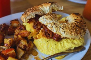 Sunny side up eggs with crispy bacon, toasted bagel, and seasoned roasted potatoes on a white plate. Perfect breakfast with fresh ingredients, hearty flavors, and savory aromas for an enjoyable morning meal.