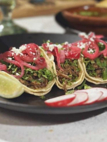 Fresh carne asada tacos on a black plate with lime wedges and radish slices, showcasing authentic Mexican street food and vibrant flavors.