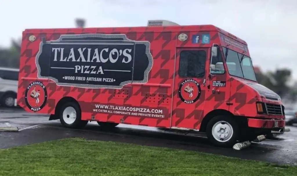 Vegan pizza food truck parked in a lot with greenery, promoting handcrafted wood-fired pizza by Tlaxiacos Pizza. Ready for catering events, offering delicious, authentic pizza on the go.
