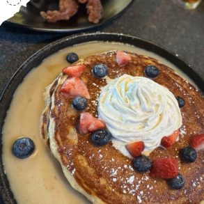 Fluffy pancake topped with whipped cream, blueberries, and strawberries served with warm syrup, as part of an international breakfast at a travel-themed restaurant.