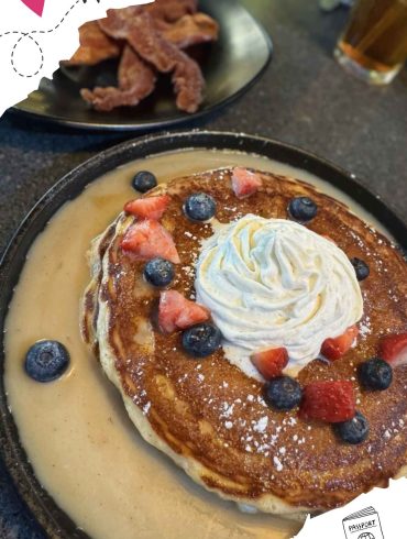 Fluffy pancake topped with whipped cream, blueberries, and strawberries served with warm syrup, as part of an international breakfast at a travel-themed restaurant.