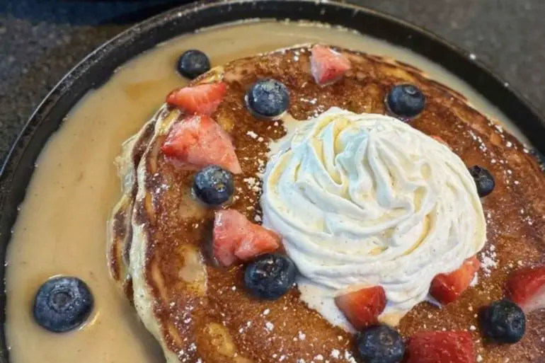 Fluffy pancake topped with whipped cream, blueberries, and strawberries served with warm syrup, as part of an international breakfast at a travel-themed restaurant.