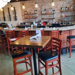 Modern restaurant interior with wooden chairs and tables, exposed brick wall, stylish pendant lighting, and a well-stocked bar area, perfect for dining and socializing.