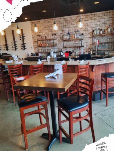 Modern restaurant interior with wooden chairs and tables, exposed brick wall, stylish pendant lighting, and a well-stocked bar area, perfect for dining and socializing.