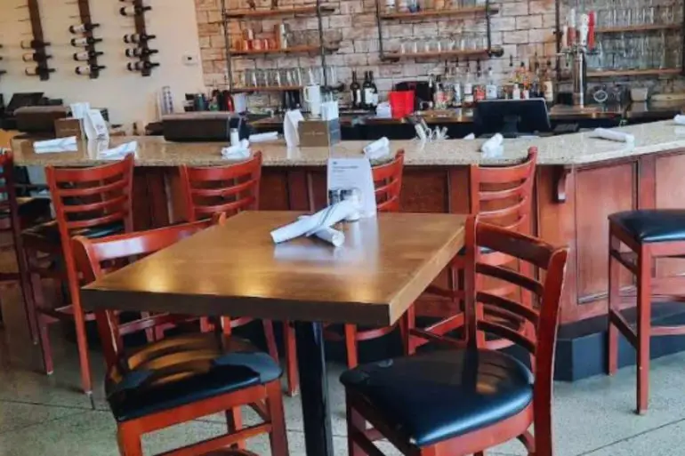 Modern restaurant interior with wooden chairs and tables, exposed brick wall, stylish pendant lighting, and a well-stocked bar area, perfect for dining and socializing.
