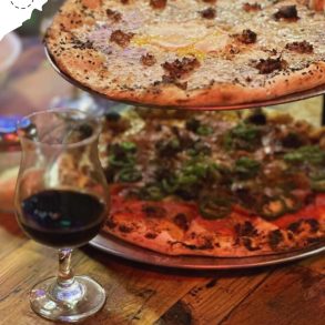 Freshly baked gourmet pizzas with melted cheese, toppings, and crispy crusts served on metal trays with a glass of red wine on a rustic wooden table.