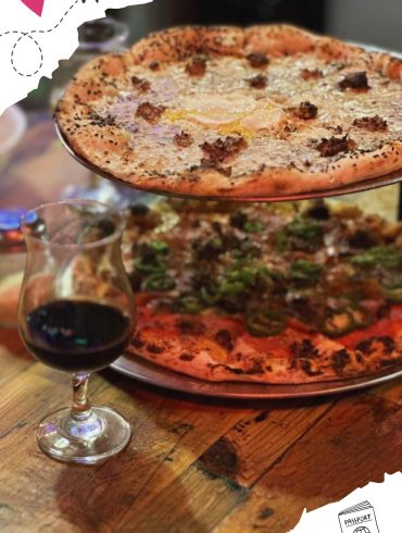 Freshly baked gourmet pizzas with melted cheese, toppings, and crispy crusts served on metal trays with a glass of red wine on a rustic wooden table.