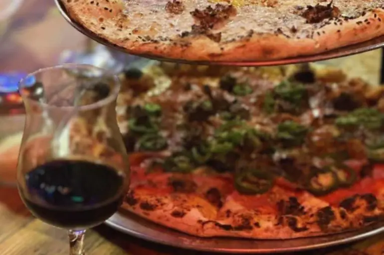 Freshly baked gourmet pizzas with melted cheese, toppings, and crispy crusts served on metal trays with a glass of red wine on a rustic wooden table.