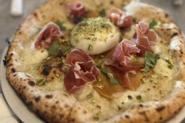 Fresh gourmet pizza with prosciutto, burrata cheese, pistachios, and basil served at a cozy pizzeria. A delicious Italian dish perfect for food lovers exploring culinary destinations.