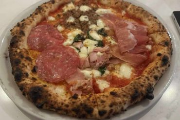 Crispy pizza topped with pepperoni, prosciutto, cheese, and fresh basil on a white plate at a restaurant in Italy.